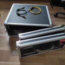 Na černé ploše transportního kufru leží černý USB kabel a žlutý audio kabel. V kufru je umístěn rackový přístroj s nápisem Fractal Audio Axe-Fx II. P…