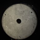 Fotografie zachycuje zadní stranu kovového reproduktorového magnetu stříbrné barvy s centrálním otvorem. Na povrchu je vyražen text: "ELECTRONIC INC"…