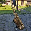 Zlatý lakovaný saxofon umístěný na černém stojanu. Nástroj má připevněný černý řemen z kůže nebo syntetického materiálu. Na povrchu nástroje nejsou v…