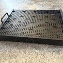 Černý kovový pedalboard s perforovaným vrchem a dvěma bočními madly. Na přední hraně je umístěn kulatý emblem s logem znázorňujícím stylizované sloup…