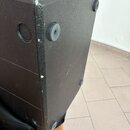Detail černé reproduktorové bedny s texturovaným povrchem. Na spodní straně jsou vidět čtyři gumové nožičky a na boku kulatý úchop. Hrany bedny vykaz…