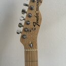 Fotografie zachycuje hlavu a horní část hmatníku elektrické kytary z přírodního světlého dřeva. Na hlavě je černý nápis Fender TELECASTER Custom. Jso…