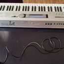 Stříbrný klavírní syntetizátor s bílými a černými klávesami. Na předním panelu je velký nápis KORG a vedle něj text TRITON Le MUSIC WORKSTATION. Přís…