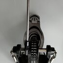 Fotografie znázorňuje černý bicí pedál s kovovými mechanismy a řetězovým přenosem. Na horní části je umístěn bílý vlněný nebo plstěný tlumicí blok na…