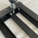 Detailní pohled na černý kovový stojan nebo držák. Součástí je nastavitelný šroub s černým plastovým knoflíkem a několika viditelnými spojovacími šro…