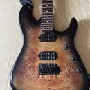 Elektrická kytara s tělem z masivu s viditnou strukturou dřeva a černým přechodem k okrajům (burst). Nástroj má dva černé humbuckery, hmatník s bílým…