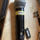 Bezdrátový mikrofon s černým tělem a stříbrnou mřížkou. Na horní části těla je nápis SM58 a logo SHURE, níže je nalepený žlutý štítek s označením 25D…