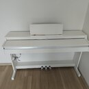 Bílé digitální piano s doprovodným stojanem a notovým pultem. Na horní liště je umístěno logo YAMAHA. Nástroj má tři pedály a bílý povrch bez viditel…