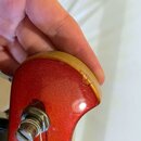 Detail hlavy kytary v červené metalizované barvě. Na horním dřevěném okraji je viditelný odlak a výrazný odštípnutý úsek materiálu. Ve spodní části j…