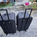 Dvě černé transportní tašky s koly a vysouvacími rukojetmi z černého plastu. Tašky jsou vyrobeny z černé textilie a vykazují mírné znečištění povrchu…