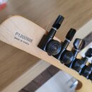 Detail hlavy kytary z přírodního dřeva s černými ladicími kolíky. Na dřevě je vytištěno sériové číslo P1200928 a nápis MADE IN KOREA. Povrch dřeva i…