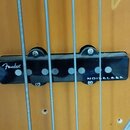 Detailní záběr na hриф basové kytary s dvěma černými snímači. Na obou snímačích je bílý nápis Fender a NOISELESS. Povrch nástroje má žlutohnědý lak,…