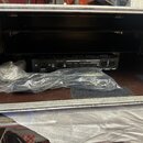 Otevřený transportní case s černým vnitřním prostorem a hnědým dnem. Uvnitř je umístěno černé zařízení s nápisy AC INPUT, PATCH BAY a RETURN 1+2 / SE…
