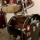 Sada červených bubnů značky Pearl s chromovaným kováním. Na basovém bubnu je černá koženka s bílým logem Pearl a několika nálepkami, včetně nápisů AL…