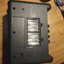 Zadní strana černého plastového elektronického zařízení s otevřeným přihrádkou pro baterie, ve které je osm AA článků. Na horní části je štítek s náp…