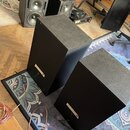 Dva černé hranaté reproduktory v popředí a další dva v pozadí. Na přední straně reproduktorů jsou vidět malé světlé štítky, z nichž na jednom je čite…