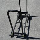 Černý transportní vozík z kovových trubek s dvěma koly. Má dvě madla, jedno v přední a jedno v zadní části konstrukce. Na spodní části jsou vidět zel…