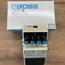 Bílý efekt pedál Boss DD-6 Digital Delay v originální krabici. Na předním panelu jsou čtyři modré oválné ovladače a nápisy CHECK, MODE (400ms, 800ms)…