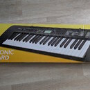 Kartonová krabice s žlutým a černým pozadím, na které je zobrazen elektronický klávesový nástroj. Na krabici jsou nápisy CASIO, ELECTRONIC KEYBOARD a…