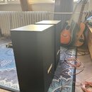Dvě černé, kvádrové reproboxové skříně umístěné na modrém vzorovaném koberci. Na boku pravého boxu je viditelný bílý štítek s textem. Z zadní strany…