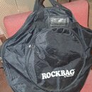 Černý textilní pouzdro (gig bag) s rukojetí a ramenním popruhem. Na přední straně je bílý nápis ROCKBAG a pod ním menší text by Warwick. Povrch mater…