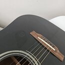 Detailní pohled na černou akustickou kytaru s tmavě hnědým dřevěným kobylníkem. Na kobylníku jsou stálé struny upevněné šesti černými kolíky. V místě…