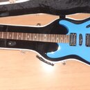Modrá elektrická kytara v tvrdém bílém kufru s černým vnitřkem. Na hlavě hmatníku je viditelné logo "Schecter". Nástroj má dva humbuckery, Tremolo sy…