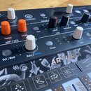 Detailní pohled na ovládací panel černého syntetizátoru s bílými a oranžovými knoflíky. Na přístroji jsou viditelné nápisy ARTURIA, ANALOG FACTORY, A…