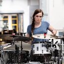 Žena s copem hraje na bicí sadu. Sada obsahuje bílé bubny s kovovými obručemi a několik činelů. Nástroje jsou umístěny v interiéru s bílými stěnami a…