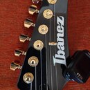 Detail hlavy černé elektrické kytary se zlatými ladicími kolíky. Na povrchu je bílý nápis Ibanez. K hlavě je připevněn černý tuner s nápisem FT20. Po…