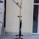 Elektrická kytara s černým korpusem a světlým hnedým hmatníkem umístěná na černém trojnožném stojanu. Kytara je držena jednou rukou za hmatník v horn…