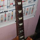 Elektrická kytara typu Les Paul v přírodním dřevěném odstínu s hnědým hmatníkem a bílými trapezoidními vložkami. Na hlavě je zlatý nápis Gibson a log…
