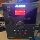 Černý elektronický bicí modul značky ALESIS, model CRIMSON II. Zařízení má digitální displej s červeným textem, kde je vidět nápis ROCK a hodnoty VOL…
