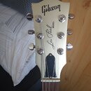 Fotografie zachycuje hlavu elektrické kytary v krémové barvě. Na povrchu jsou čitelné nápisy „Gibson“, „Les Paul“ a menší text „MODEL“. Nástroj má še…