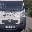 Na fotografii je bílý dodávkový vůz s šedým nárazníkem, který stojí na asfaltové cestě. Na kapotě je černý nápis v dekorativním písmu „motörhit“. Vůz…