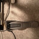 Černý sustain pedál značky Roland s kovovou stoupací plochou. Pedál je připojen k černému kabelu a položen na šedém povrchu vedle černého stojanu. Po…