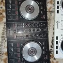 Černý DJ kontrolér s dvěma stříbřistými jog-koly a centrální mixážní sekcí s posuvníky a knoflíky. Na zařízení jsou vidět nápisy Pioneer DJ, serato a…
