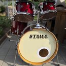 Vícedílná bicí souprava v tmavě červené barvě s chromovaným kováním. Na přední straně basového bubnu je černý nápis TAMA®. Horní tommy mají na spodní…