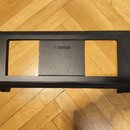 Černý plastový stojan nebo podstavec s obdélníkovým tvarem a dvěma výřezy. Uprostřed je vyraženo logo a nápis YAMAHA. Povrch vykazuje mírné známky op…