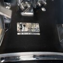 Detailní pohled na černý povrch bubnu s kovovými komponenty. Na korpusu je připevněna stříbrná tabulky s nápisem SONOR a textem Made in Germany, pod…