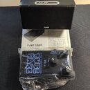 Černý malý kytarový zesilovač v plastovém obalu, doprovázený krabicí a dokumentací. Na zařízení je bílý nápis TUBE CAKE a text GUITAR POWER AMPLIFIER…