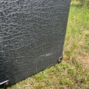 Detailní záběr na černý povrch s texturou imitující kůži, pravděpodobně z pouzdra nebo zesilovače. Povrch vykazuje mírné oděrky a světlé šmouhy v dol…