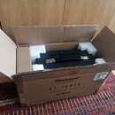 V otevřeném kartonovém boxu s bílými polystyrenovými výplněmi je umístěn černý kytarový kombozesilovač s černou rukojetí a kovovými detaily. Na předn…