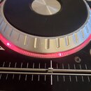 Detailní pohled na DJ kontroler s velkým černým jog-wheelem a stříbrným rýhovaným okrajem. Pod okrajem kola je vidět červené podsvícení a pod ním se…