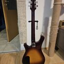 Elektrická kytara s trupem v tmavě hnědé a žluté barvě (sunburst) umístěná na černém stojanu. Nástroj je zobrazen z pozadí, kde je v dolní části vidě…