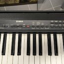 Černý digitální klavír s bílými a černými klávesami. Na ovládacím panelu je uprostřed bílý nápis YAMAHA a v pravém dolním rohu označení ELECTRONIC PI…