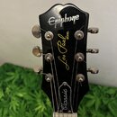 Fotografie zachycuje hlavu černé elektrické kytary. V horní části je bílý nápis Epiphone a uprostřed žlutý nápis Les Paul MODEL. Dole se nachází bílý…
