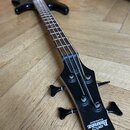 Černá elektrická basová kytara s hnedým hmatníkem leží na dřevěné podlaze. Na hlavě nástroje je bílý nápis Ibanez a GIO s doplňkem SOUNDGEAR. Na zadn…