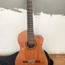 Akustická kytara s vyřezem v korpusu, provedená v přirozené dřevěné barvě. Na hmatníku je vidět štítek s nápisem "Classical Guitar". Nástroj stojí op…