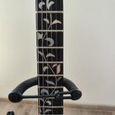 Detail hmatníku černé elektrické kytary s ozdobnou vložkou ve tvaru rostlinného ornamentu z bílého materiálu. Na hmatníku jsou vidět kovové pražce a…